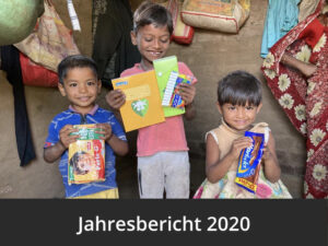 Zwei Jungen und ein kleines Mädchen lächeln. Sie stehen in ihrem Zuhause und haben Lehrmittel und, Schokolade und Stifte in den Händen. Diese wurden im Rahmen der Corona Nothilfe von Mudita School, dem Projekt von Bharat Wankhade aus Indien, verteilt.